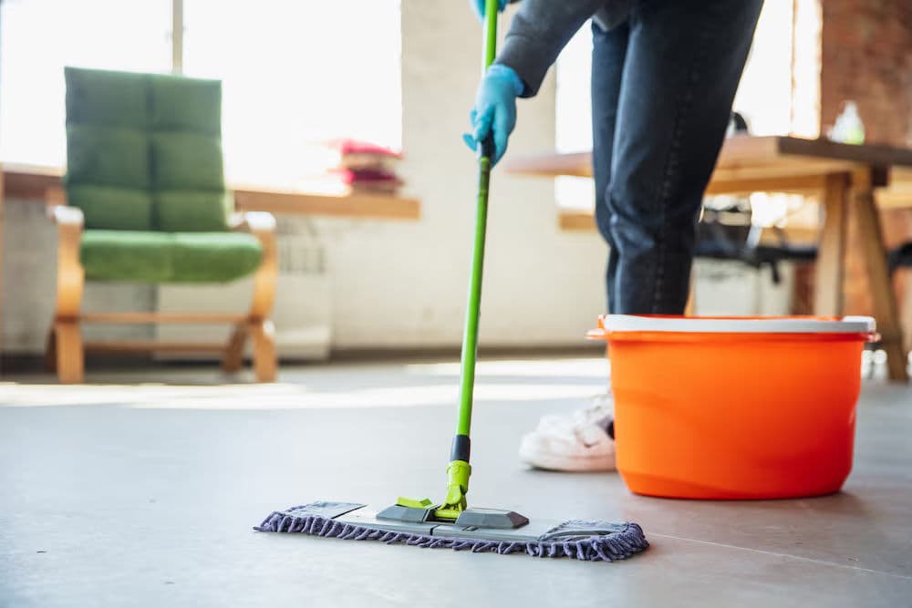 Person mopping a floor with a green mop and orange bucket in a bright, modern space.